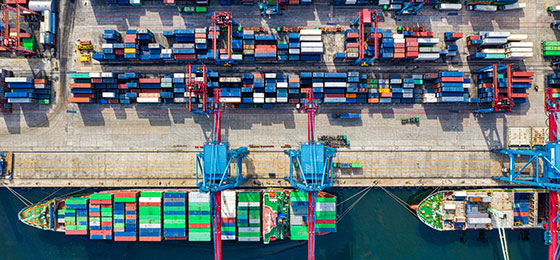 Vogelperspektive auf ein Frachtschiff mit Containern an einem Hafen mit Containerkränen und weiteren Seefracht-Containern auf de