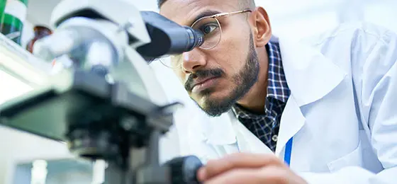 Wissenschaftler mit Mikroskop im Labor. Sein Gesich ist halb vom Mikroskop verdeckt.