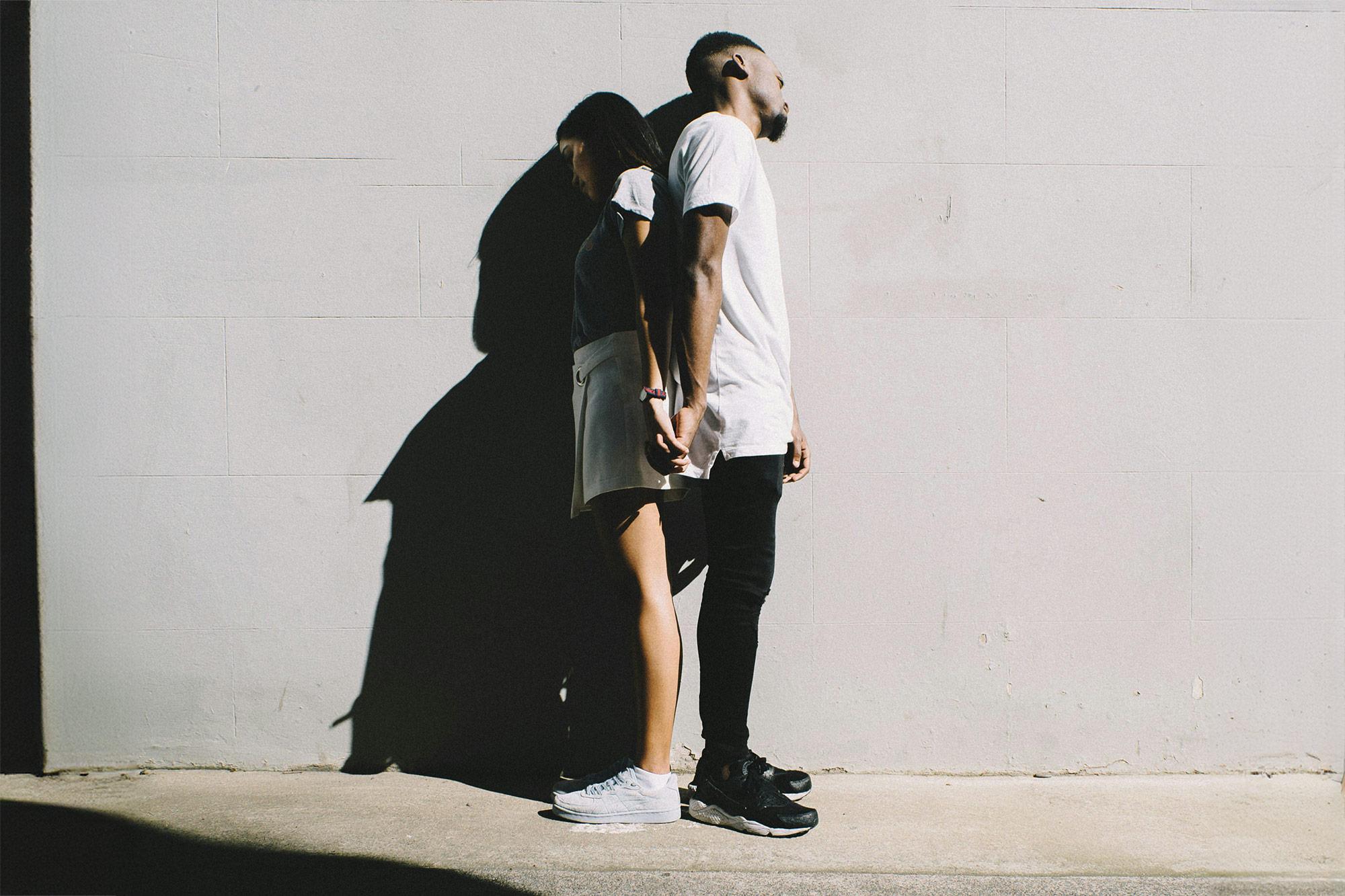 Ein junges Paar lehnt händchenhaltend, aber Rücken an Rücken an einer Wand. 